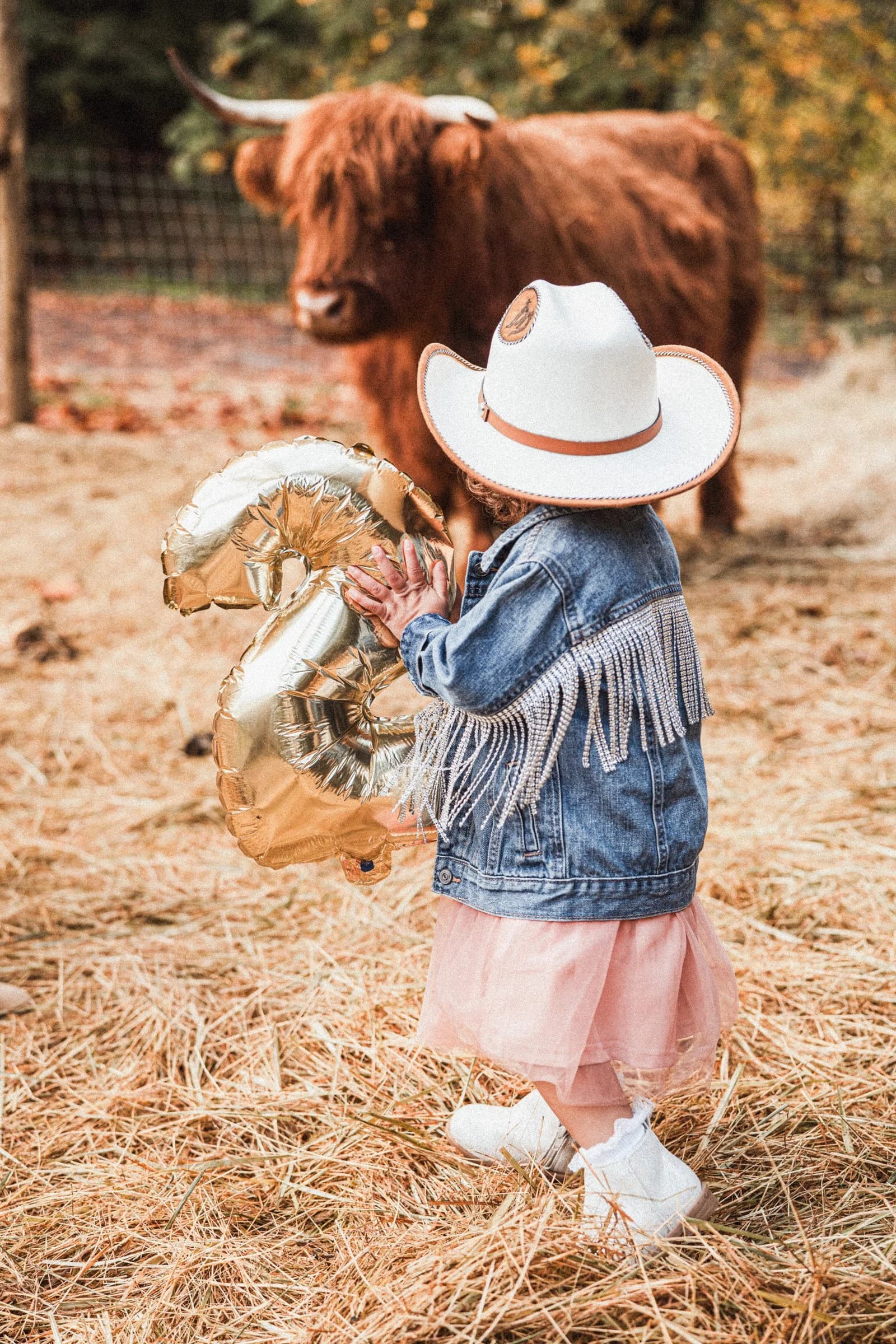 A child in a white cowboy hat and fringed denim jacket holds a gold 3 balloon beside a Highland cow
