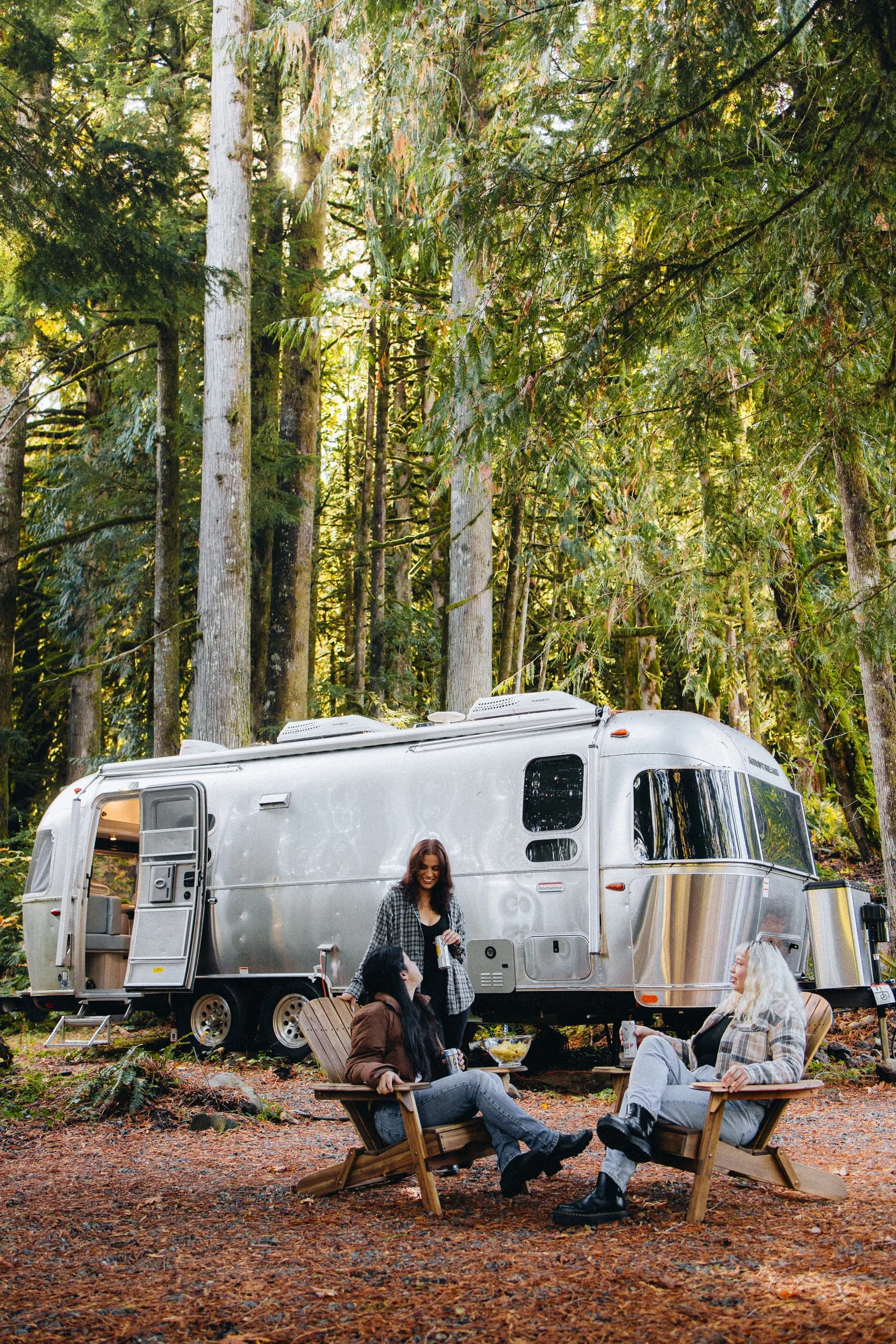 Guests in Adirondack chairs beside the Airstream at the Camp, under tall trees