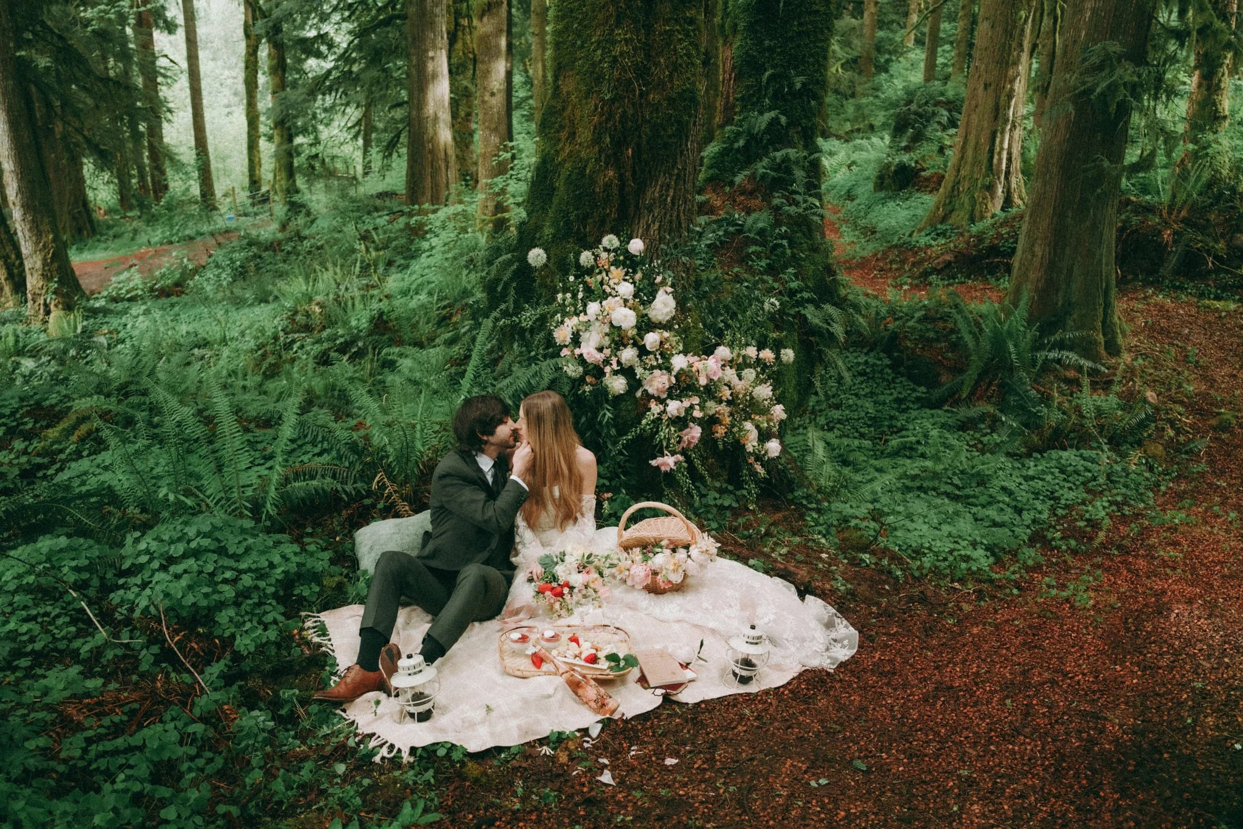 Two people in wedding clothes picnic on a blanket under the trees beside a bouquet