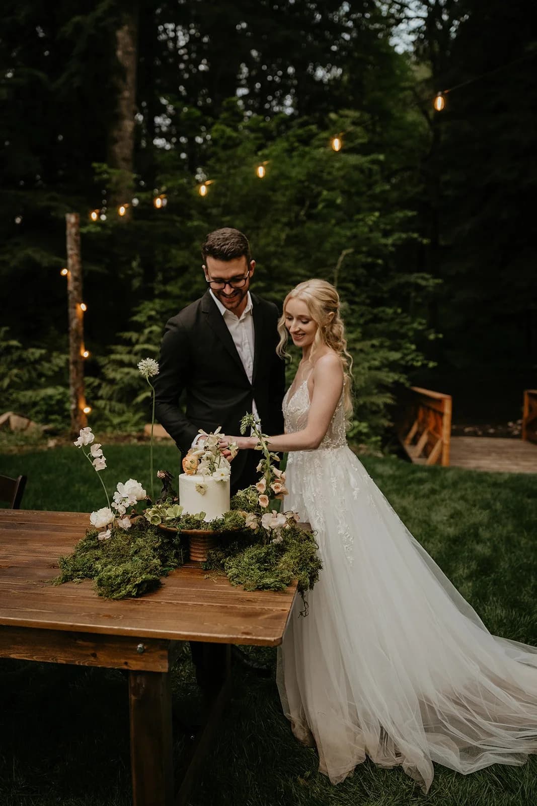 Two people in wedding clothes cut a cake at a long wooden table among the trees