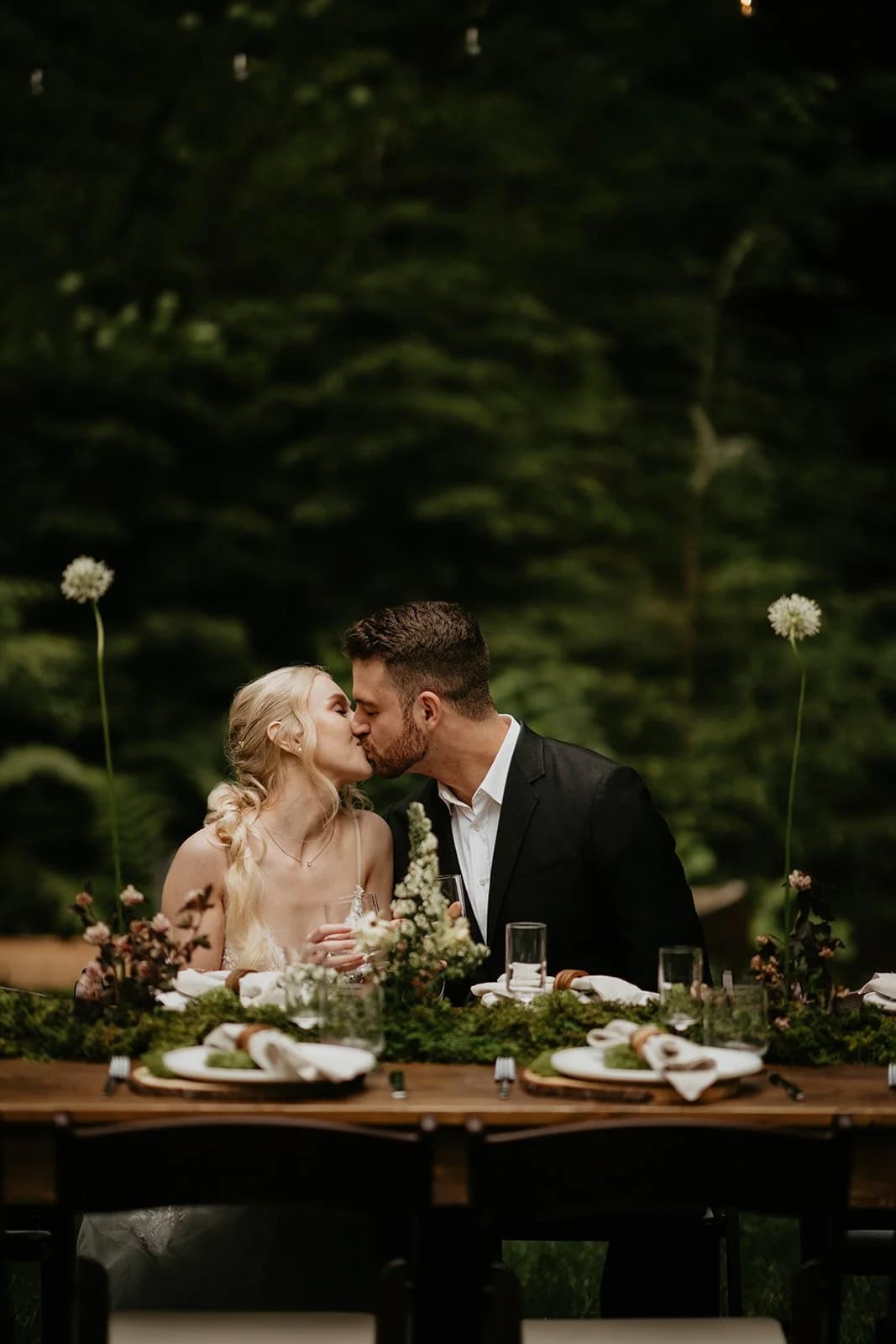 Two people in wedding clothes kiss over a table set with moss and flowers