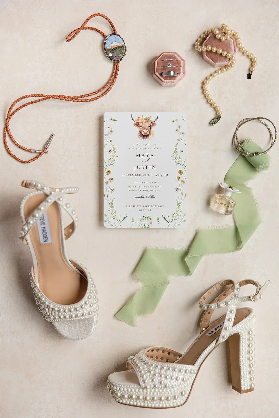 Maya and Justin's wedding invitation, topped with a painted Highland cow in a flower crown, among pearl shoes and a bolo tie
