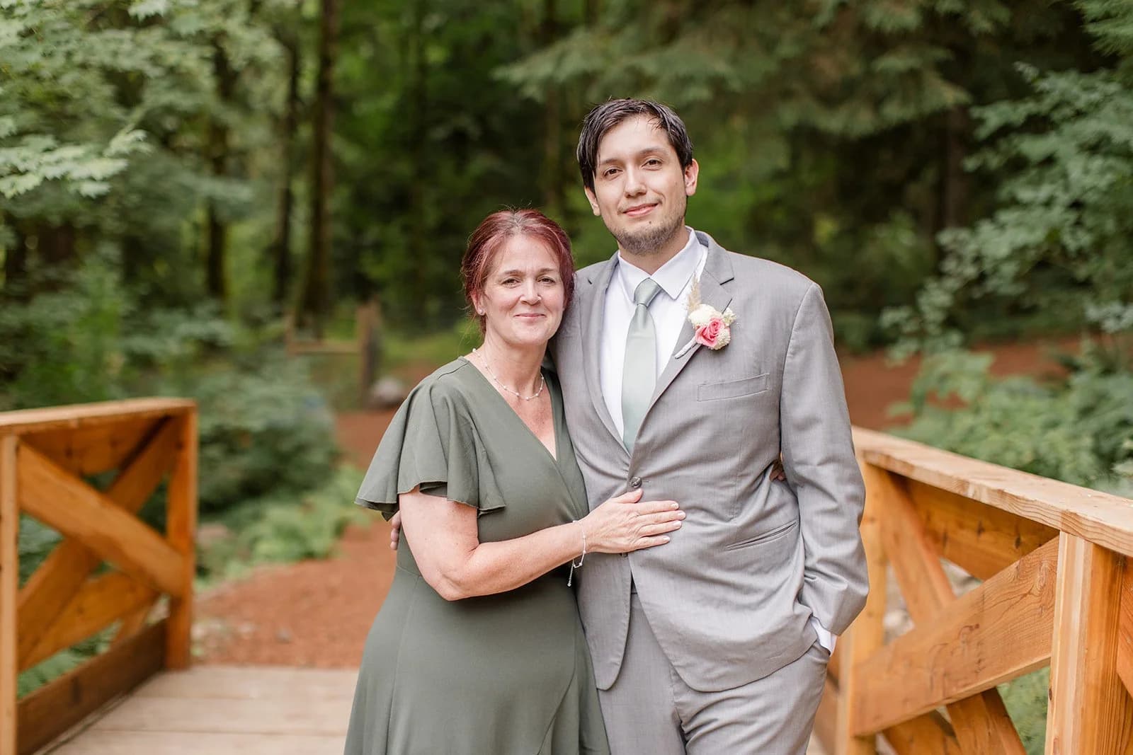 A man in a grey suit with a rose boutonniere stands arm in arm with a woman in a sage dress on a wooden footbridge