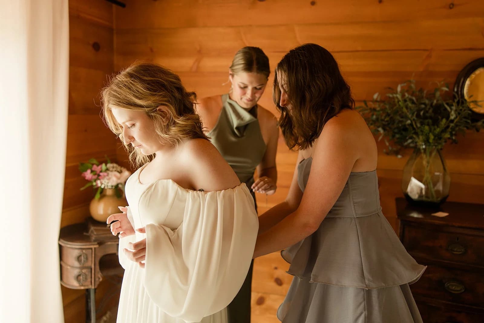 Two women in sage dresses fasten the bride's gown in a warm cedar-walled room