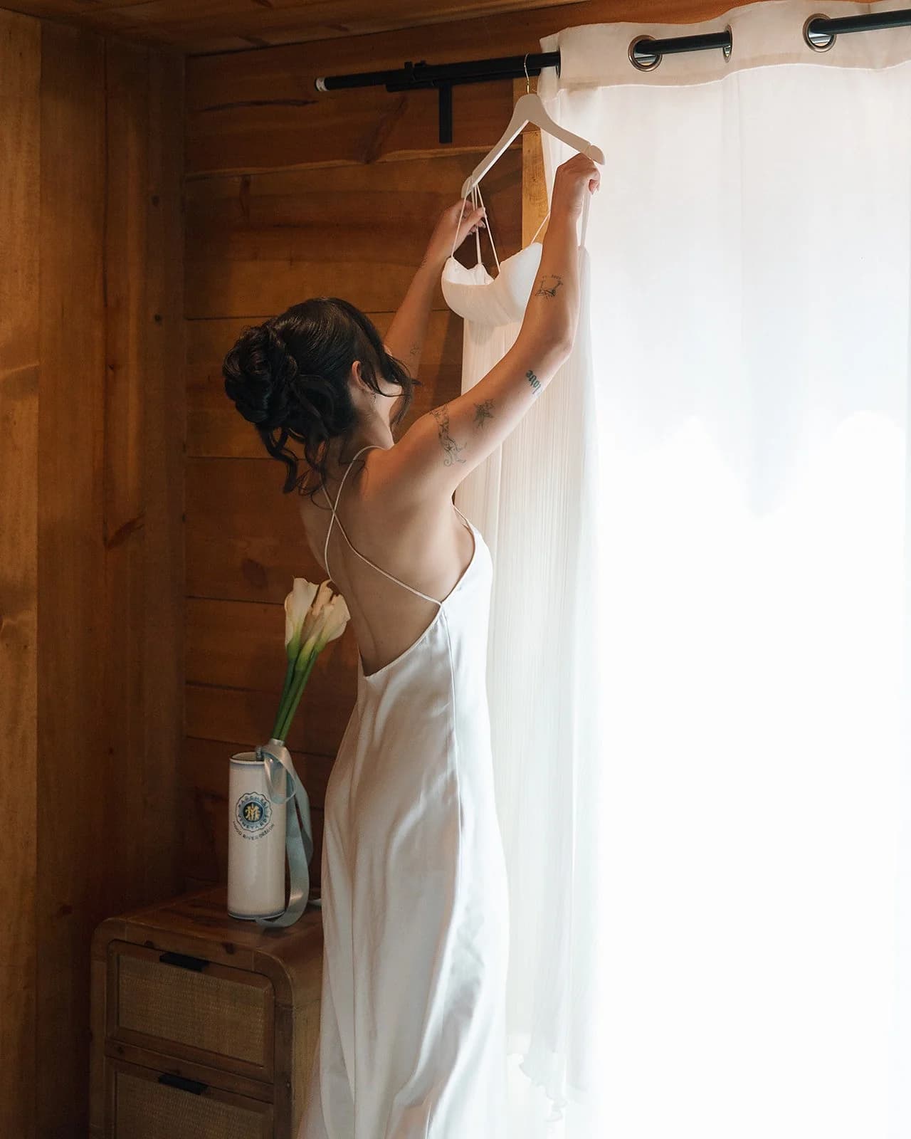 A bride in a slip dress reaches up to her gown hanging by a curtained window