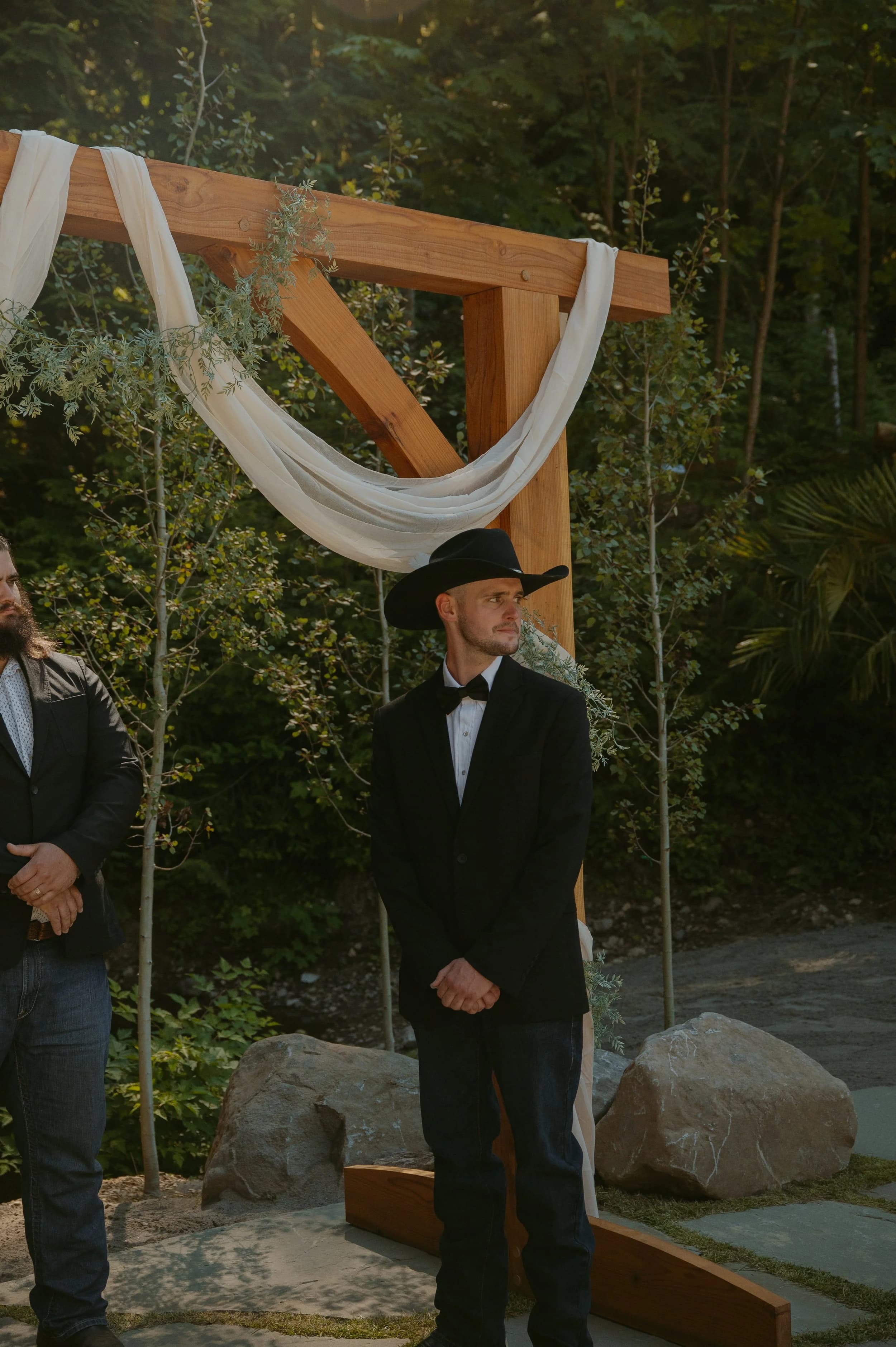 A groom in a black cowboy hat waits beside a timber arch draped in cream fabric
