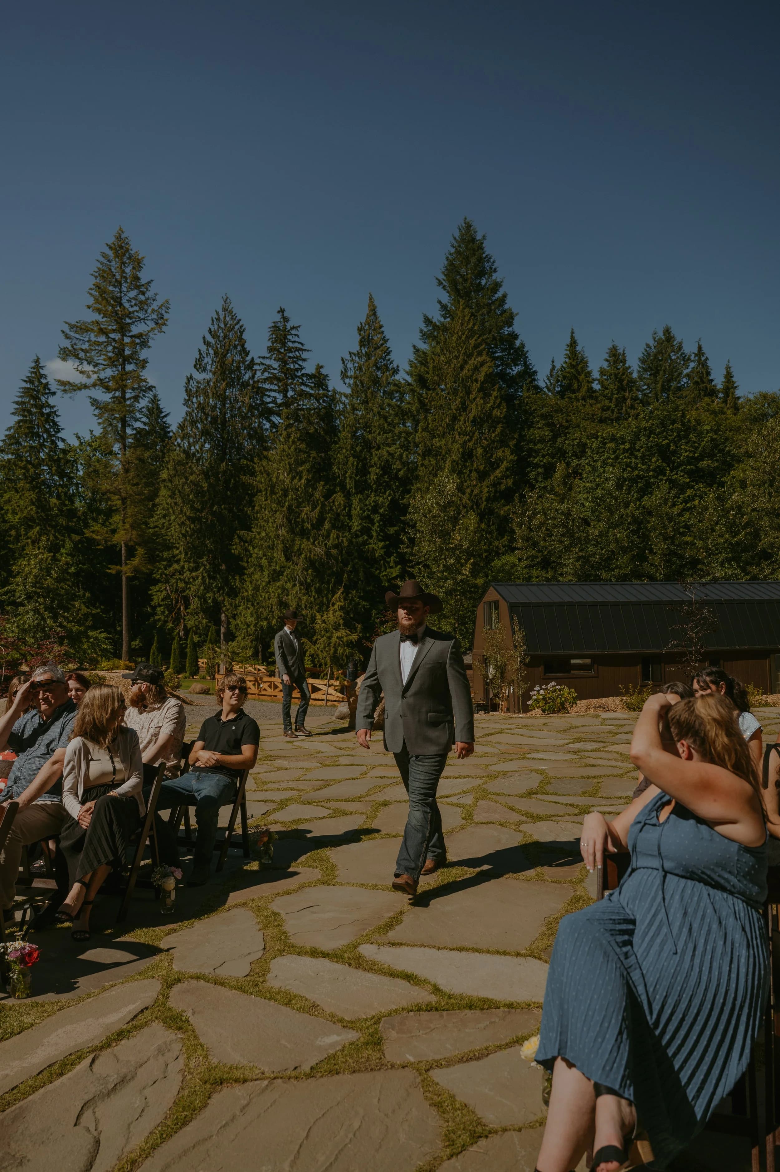 Guests seated on a flagstone patio as a man in a grey suit walks the aisle