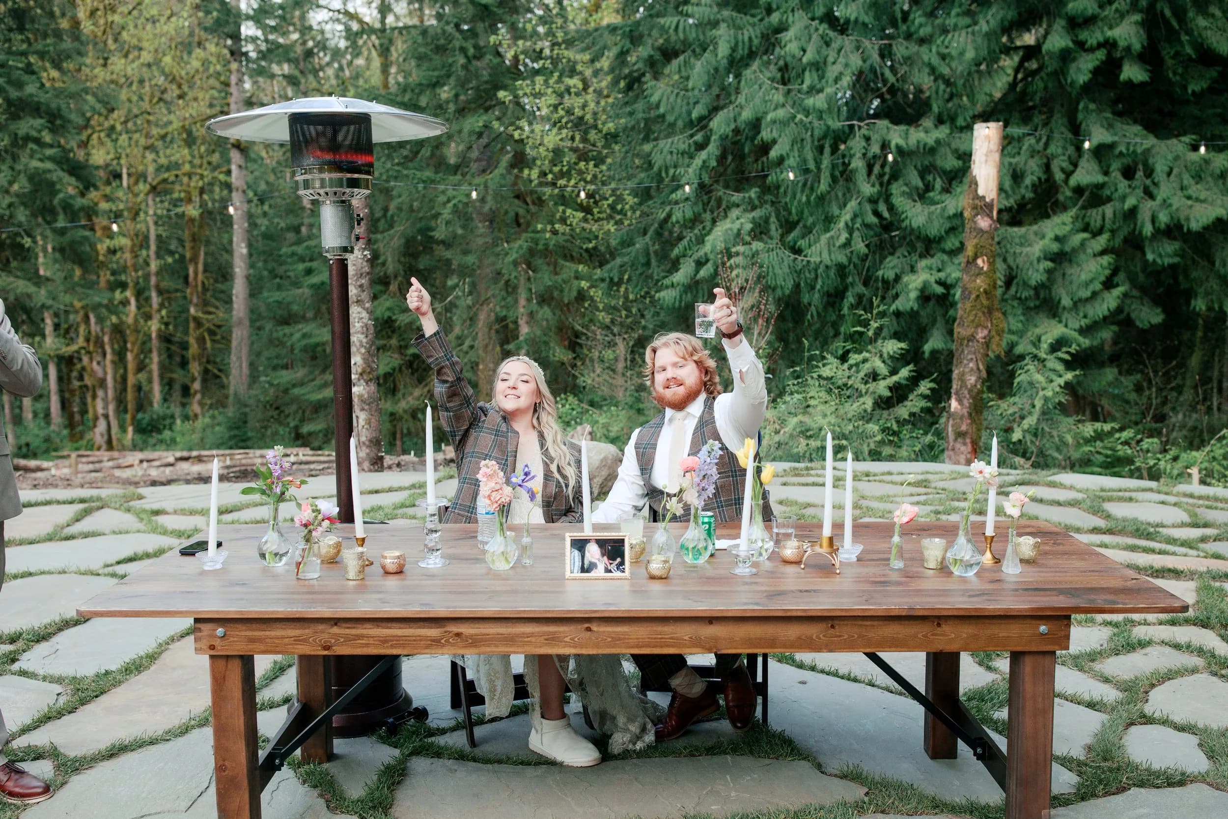 A couple at a long table set with candles and glasses, one arm raised