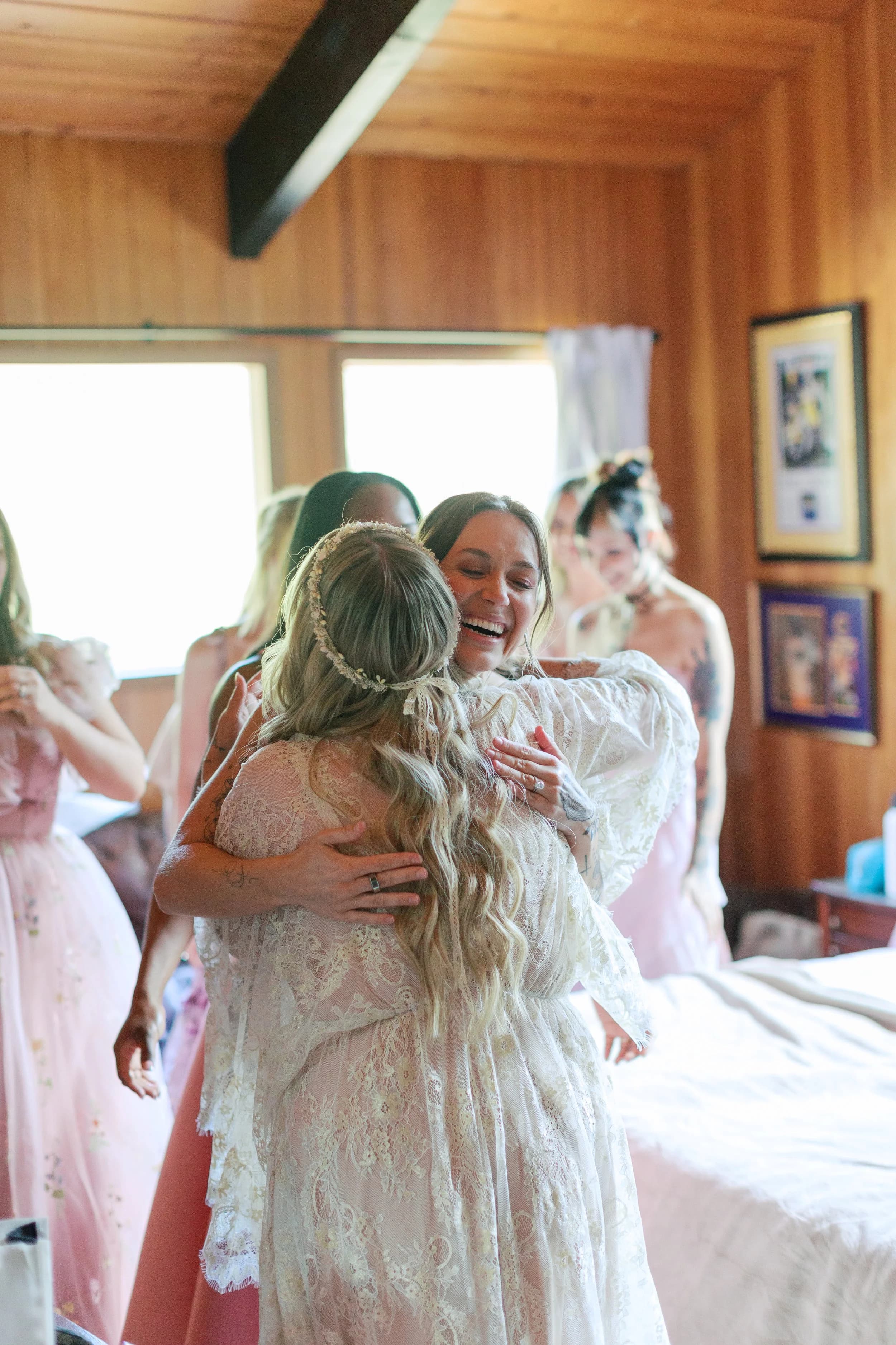 Bridesmaids in pink and cream embrace the bride in a warm cedar-walled room