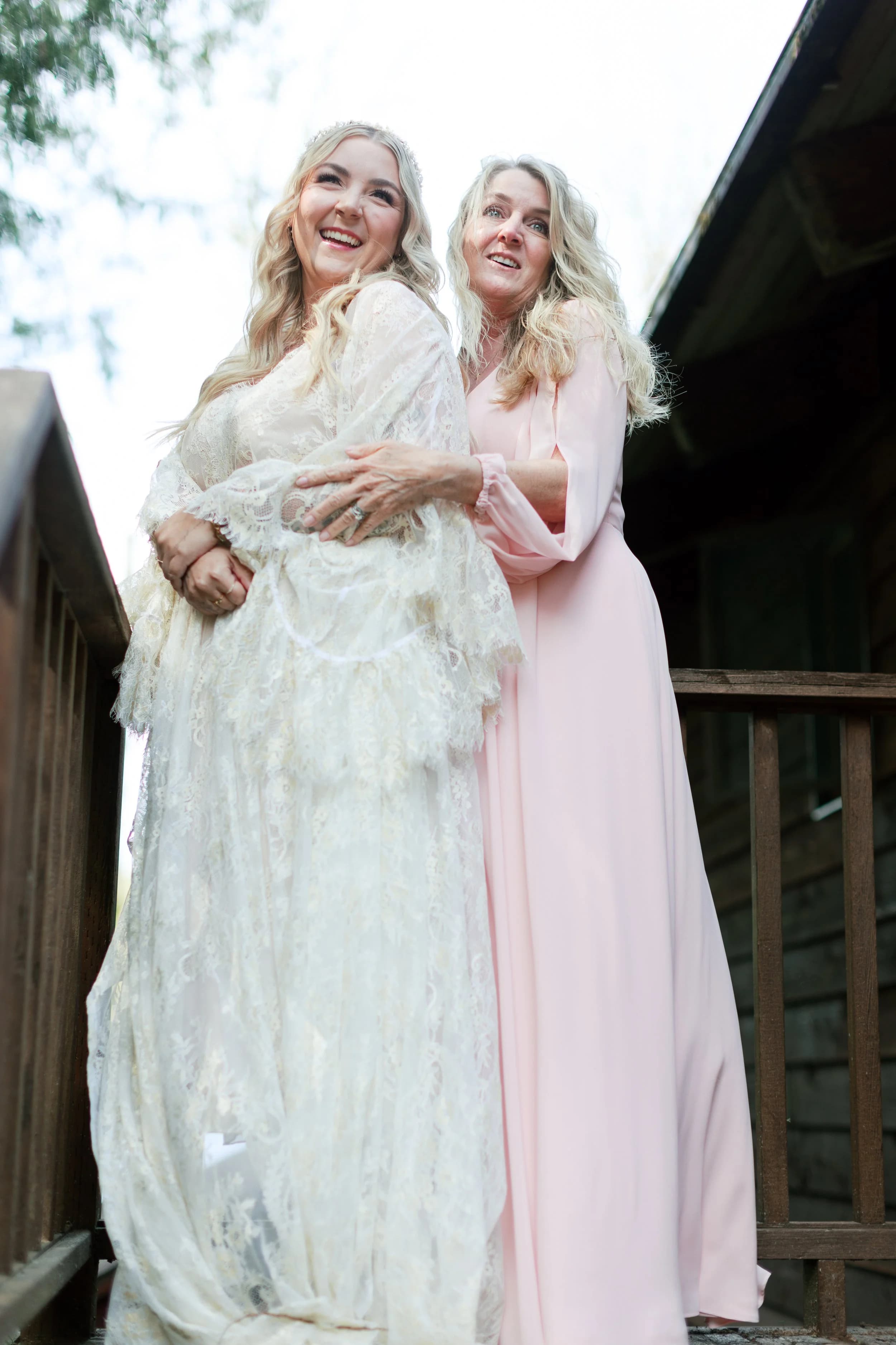 The bride, in a white shawl, laughs with a bridesmaid in pink on a wooden deck